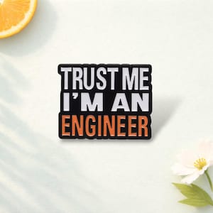 May include: A rectangular enamel pin with a black background and white text that reads TRUST ME I'M AN, followed by the word ENGINEER in orange capital letters. The pin features a bold, sans-serif font and a clean, defined border design.