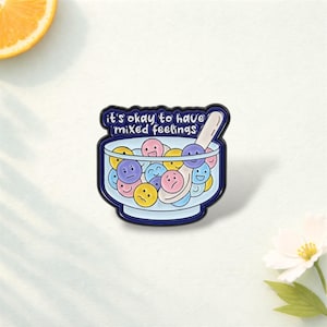 May include: Enamel pin featuring a bowl of colorful smiley faces with various expressions, and a spoon. The pin has the text "it's okay to have mixed feelings" in a blue banner above the bowl. The pin is on a white surface.