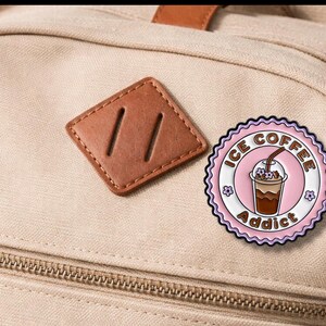May include: A round, pink and white enamel pin with the words "ICE COFFEE Addict" encircling an illustration of an iced coffee drink. The pin is affixed to a beige backpack.