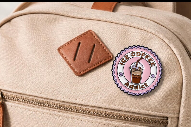 May include: A round, pink and white enamel pin with the words "ICE COFFEE Addict" encircling an illustration of an iced coffee drink. The pin is affixed to a beige backpack.