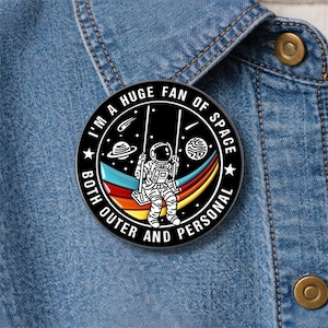 以下が含まれることがあります： 「I'M A HUGE FAN OF SPACE BOTH OUTER AND PERSONAL」の文字が入った、丸い黒いエナメルピン。虹色の背景、惑星、星を背景にブランコに乗る宇宙飛行士が描かれています。デニムジャケットにピンが留められています。