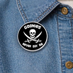 Könnte beinhalten: Ein runder, schwarzer Button mit dem Text "GOONIES" über einem Totenkopf und gekreuzten Schwertern sowie den Worten "NEVER SAY DIE" darunter. Der Button befindet sich auf einem blauen Jeanshintergrund.