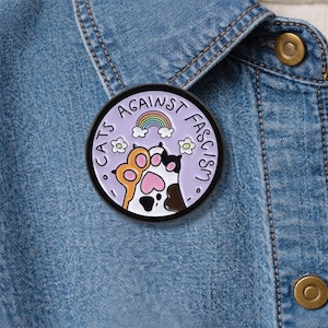 Op de afbeelding: Ronde emaille pin met de tekst "CATS AGAINST FASCISM" in zwart. De pin toont een regenboog, wolken, bloemen en kattenpoten in oranje, zwart en wit op een lavendel achtergrond. Op een spijkerjasje.
