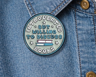 May include: A round enamel pin with the text "Introverted but willing to discuss books." The pin features a light blue background with white text and star accents. Two stacked books are in the center. The pin is on a blue denim jacket.