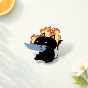 Può includere: Una spilla smaltata a forma di gatto nero, raffigurante un felino in stile cartoon con orecchie a punta e coda, che tiene in mano un portatile azzurro. Dietro il gatto c'è un disegno stilizzato di fiamme arancioni e gialle. Occhi gialli.