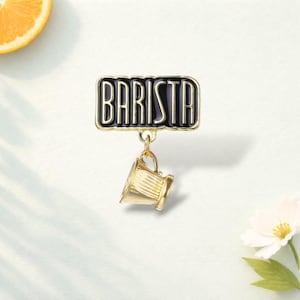 May include: A gold-toned metal pin featuring a rectangular black enamel plaque with the word BARISTA in raised gold lettering. A small, dangling gold-toned coffee cup charm is attached to the bottom of the plaque by a circular metal jump ring.