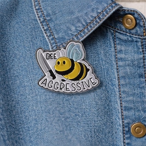 May include: A yellow and black enamel pin featuring a cartoon bee holding a knife. The pin has the words "BEE AGGRESSIVE" in a speech bubble design. The pin is set against a blue denim jacket.