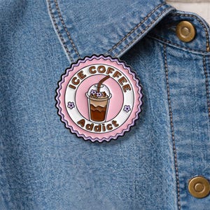 May include: A round, pink enamel pin with the words "ICE COFFEE Addict" encircling a graphic of an iced coffee drink. The pin has a scalloped edge and is decorated with small flower accents. The pin is attached to a denim jacket.