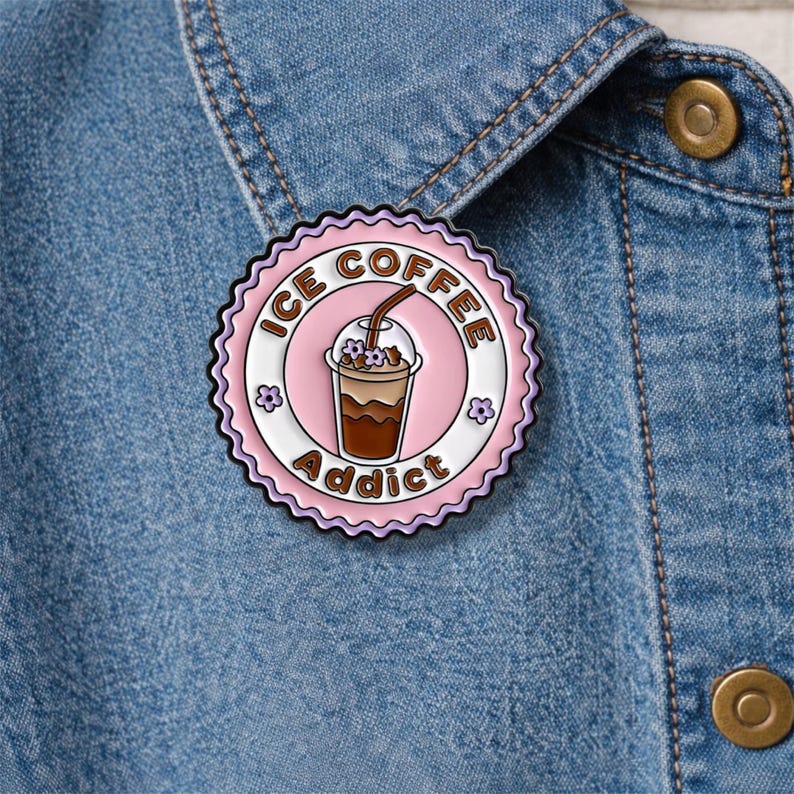 May include: A round, pink enamel pin with the words "ICE COFFEE Addict" encircling a graphic of an iced coffee drink. The pin has a scalloped edge and is decorated with small flower accents. The pin is attached to a denim jacket.