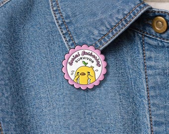 May include: A pink and white enamel pin with the text "Social Gathering Survivor" encircling a yellow chick with tears. The pin is attached to a blue denim jacket.