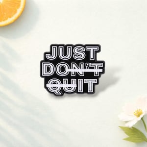 Può includere: Una spilla in smalto nero con il testo bianco JUST DON'T QUIT. Le parole DON'T e QUIT sono ciascuna barrate da una linea orizzontale bianca, lasciando la parola JUST come unico testo visibile che non è barrato.