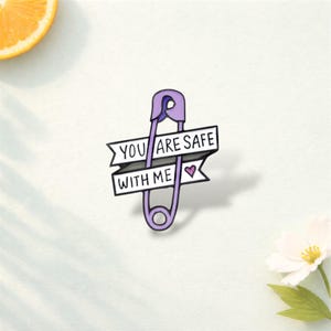 May include: A lilac safety pin with the text "YOU ARE SAFE WITH ME" in black and white. A small purple heart is next to the text. The pin is on a white background.