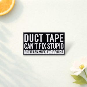 Puede incluir: Pin rectangular negro con texto blanco: "DUCT TAPE CAN'T FIX STUPID BUT IT CAN MUFFLE THE SOUND". Sobre una superficie blanca, con una rodaja de naranja y una flor blanca.