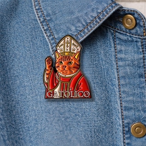 Puede incluir: Un colorido pin de esmalte con un gato de dibujos animados vestido como obispo católico, con mitra y vestiduras rojas. El pin tiene la palabra "GATOLICO" en la parte inferior. El pin está sujeto a una chaqueta vaquera.