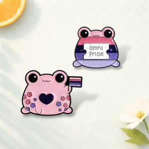 Puede incluir: Dos pines de esmalte con ranas de dibujos animados. Una rana sostiene una bandera con un diseño de rayas, y la otra sostiene un letrero que dice "Omni Pride". Las ranas son rosas con ojos y acentos negros.
