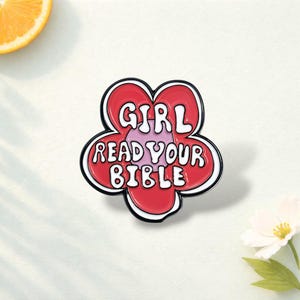 Può includere: Una spilla in smalto rosso e bianco a forma di fiore con le parole "GIRL READ YOUR BIBLE" in lettere bianche. La spilla ha un contorno nero e un centro rosa chiaro.