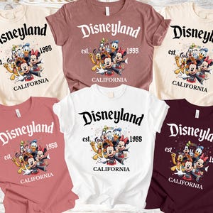 May include: A collection of short-sleeved t-shirts in various colors, each featuring the Disneyland logo with the text "est. 1955 CALIFORNIA" and a graphic of Disney characters. The shirts are in cream, mauve, white, and burgundy.