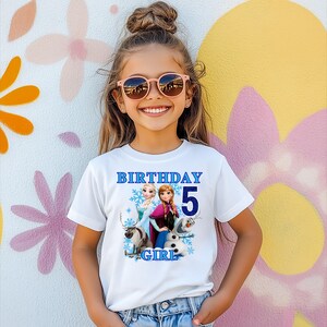 Może przedstawiać: Biała koszulka z niebieskim napisem "BIRTHAY 5 GIRL", przedstawiająca postacie z filmu Kraina Lodu. Koszulka jest noszona przez młodą osobę w r&oacute;żowych okularach przeciwsłonecznych, uśmiechniętą przed kolorową ścianą.