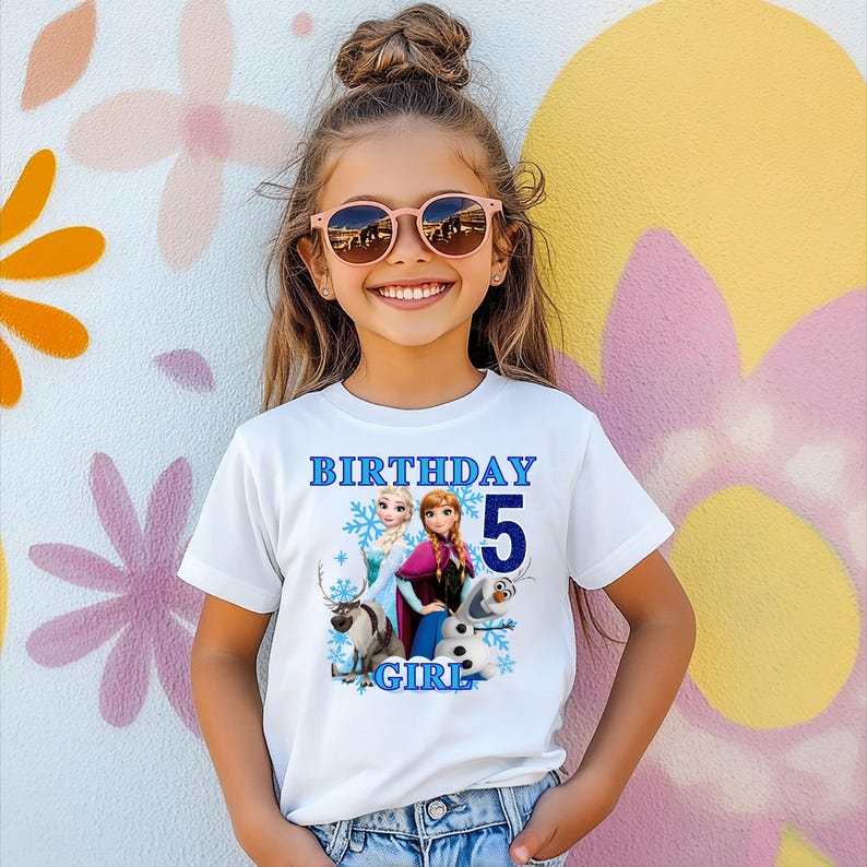 Może przedstawiać: Biała koszulka z niebieskim napisem "BIRTHAY 5 GIRL", przedstawiająca postacie z filmu Kraina Lodu. Koszulka jest noszona przez młodą osobę w r&oacute;żowych okularach przeciwsłonecznych, uśmiechniętą przed kolorową ścianą.