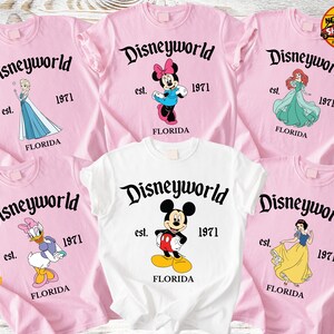 Op de afbeelding: Roze en witte T-shirts met Disney-personages en de tekst "Disneyworld est. 1971 Florida." Elk shirt heeft een ander personage: Elsa, Minnie Mouse, Ariel, Katrien Duck, Mickey Mouse en Sneeuwwitje.
