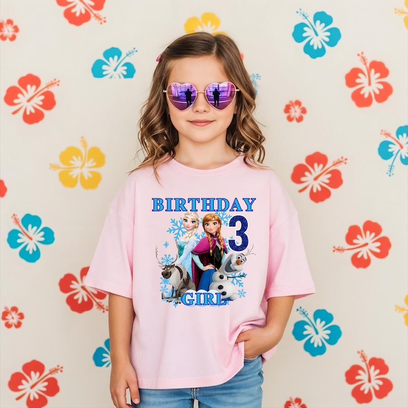 Może przedstawiać: Dziewczynka w r&oacute;żowej koszulce z grafiką z filmu Kraina Lodu i niebieskim napisem "BIRTHDAY 3 GIRL". Ma r&oacute;wnież okulary przeciwsłoneczne w kształcie serca. Tło przedstawia wz&oacute;r kwiatowy.