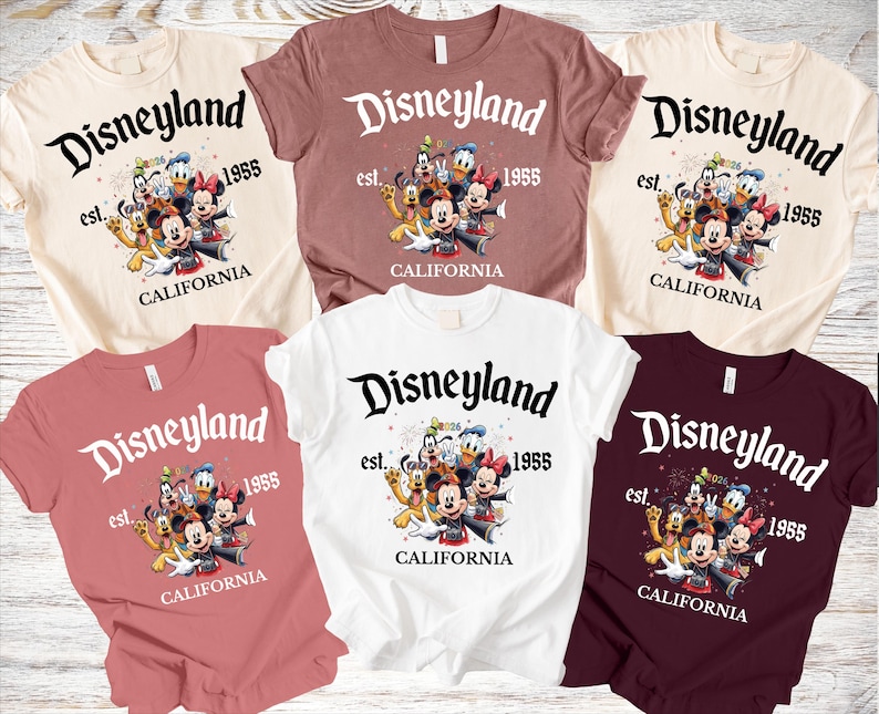 May include: A collection of short-sleeved t-shirts in various colors, each featuring the Disneyland logo with the text "est. 1955 CALIFORNIA" and a graphic of Disney characters. The shirts are in cream, mauve, white, and burgundy.