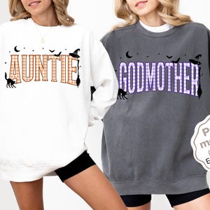 May include: A white sweatshirt with the orange gingham text AUNTIE and a dark gray sweatshirt with the purple gingham text GODMOTHER. Both feature black graphics of a crescent moon, stars, bats, a witch hat, a broom, and a black cat.