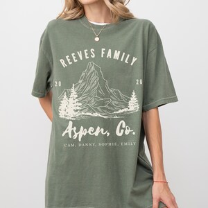 Pu&ograve; includere: T-shirt verde oliva con grafica di montagna e la scritta "REEVES FAMILY Aspen, Co.". La maglietta include anche i nomi "CAM, DANNY, SOPHIE, EMILY". La maglietta ha una vestibilit&agrave; rilassata.