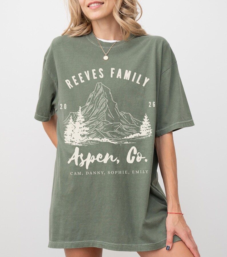 Pu&ograve; includere: T-shirt verde oliva con grafica di montagna e la scritta "REEVES FAMILY Aspen, Co.". La maglietta include anche i nomi "CAM, DANNY, SOPHIE, EMILY". La maglietta ha una vestibilit&agrave; rilassata.