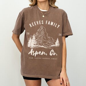 Pu&ograve; includere: T-shirt marrone con la scritta "REEVES FAMILY" sopra una grafica di montagna e "Aspen, Co." sotto. La maglietta include anche i numeri "20" e "26" e i nomi "CAM, DANNY, SOPHIE, EMILY".