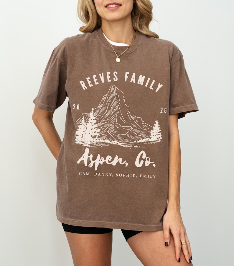 Pu&ograve; includere: T-shirt marrone con la scritta "REEVES FAMILY" sopra una grafica di montagna e "Aspen, Co." sotto. La maglietta include anche i numeri "20" e "26" e i nomi "CAM, DANNY, SOPHIE, EMILY".