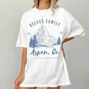 Pu&ograve; includere: T-shirt bianca con grafica di montagna blu. Il testo sulla maglietta recita "REEVES FAMILY 2026 Aspen, Co." e include nomi. Il design presenta una catena montuosa e pini.