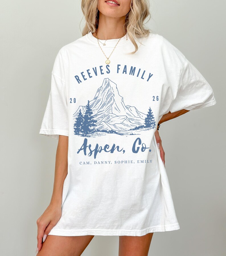 Pu&ograve; includere: T-shirt bianca con grafica di montagna blu. Il testo sulla maglietta recita "REEVES FAMILY 2026 Aspen, Co." e include nomi. Il design presenta una catena montuosa e pini.
