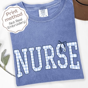 May include: A blue short-sleeved t-shirt with a vintage wash finish. The front features the word NURSE in large block letters with a light blue and white gingham pattern. A small dark blue bow graphic sits atop the letter S. A label reads Comfort Colors.