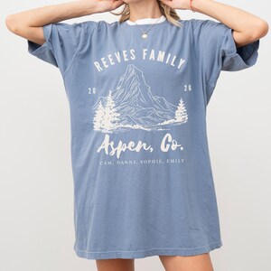 Pu&ograve; includere: Una maglietta blu con testo bianco e una grafica di montagna. La maglietta recita "Reeves Family" sopra un'illustrazione di montagna, con "Aspen, Co." sotto. I nomi Cam, Danny, Sophie ed Emily sono elencati. La maglietta ha un colletto bianco.