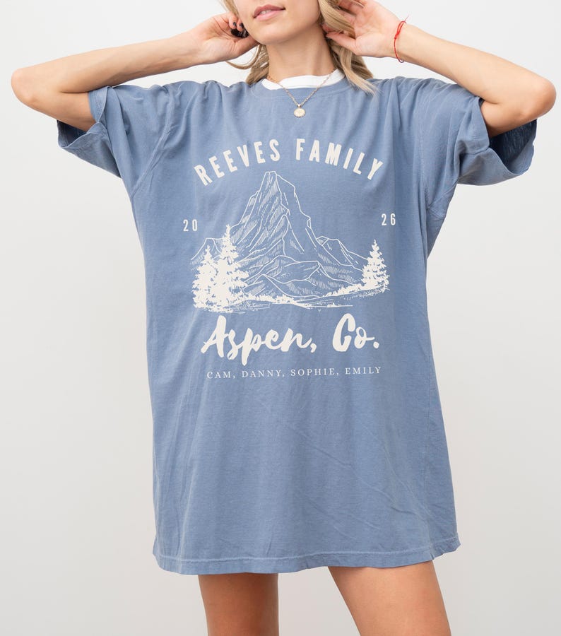 Pu&ograve; includere: Una maglietta blu con testo bianco e una grafica di montagna. La maglietta recita "Reeves Family" sopra un'illustrazione di montagna, con "Aspen, Co." sotto. I nomi Cam, Danny, Sophie ed Emily sono elencati. La maglietta ha un colletto bianco.