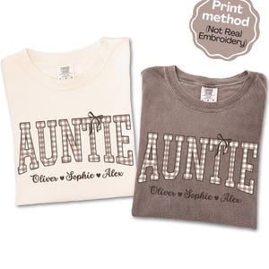May include: Two crewneck t-shirts, one cream and one dark gray, featuring the word AUNTIE in a brown and white gingham pattern with a small bow graphic. Below the text, the names Oliver, Sophie, and Alex are printed with heart icons.