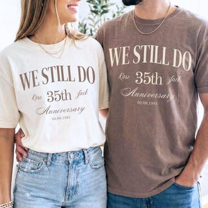 May include: Two people wearing matching t-shirts. The shirts are cream and brown, each with the text "WE STILL DO" above "Rose 35th Jack Anniversary 02.08.1991" in a script font. They are wearing blue jeans.