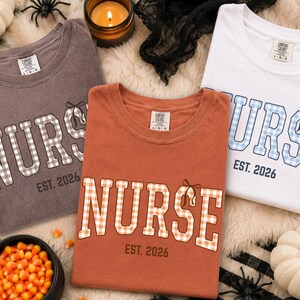 May include: Three crewneck t-shirts in charcoal, burnt orange, and white. Each shirt features the word NURSE in a gingham-patterned block font with a small bow graphic on the letter E, accompanied by the text EST. 2026 printed in a dark color.