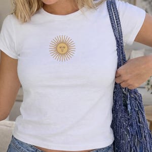 May include: A white short-sleeved t-shirt featuring a centered graphic of a yellow sun with a human-like face and radiating rays. The sun design is rendered in thin brown lines, providing a clear contrast against the plain white fabric of the shirt.