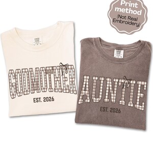 May include: Two crewneck t-shirts, one cream and one dark gray, featuring brown and white gingham block lettering. The cream shirt reads GODMOTHER EST. 2026 and the gray shirt reads AUNTIE EST. 2026, both with a small bow graphic. Print method (Not Real Embroidery).