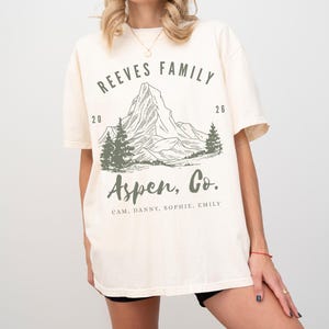 Pu&ograve; includere: T-shirt color crema con grafica di montagna e la scritta "REEVES FAMILY Aspen, Co." in verde. Il design include una catena montuosa, alberi e i nomi CAM, DANNY, SOPHIE e EMILY. La maglietta ha maniche corte.
