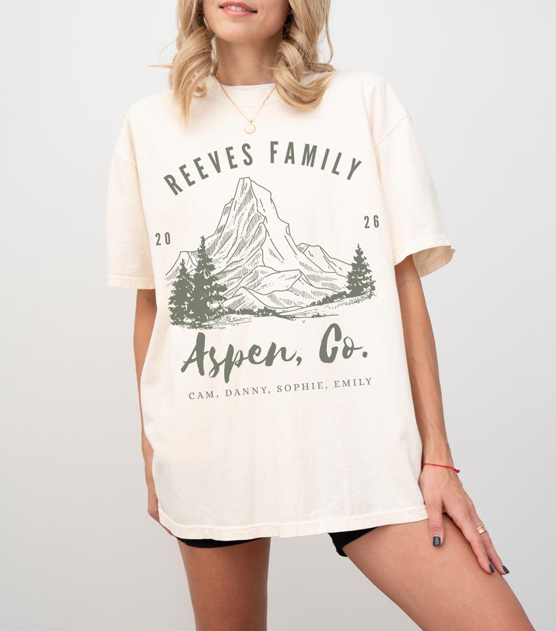Pu&ograve; includere: T-shirt color crema con grafica di montagna e la scritta "REEVES FAMILY Aspen, Co." in verde. Il design include una catena montuosa, alberi e i nomi CAM, DANNY, SOPHIE e EMILY. La maglietta ha maniche corte.