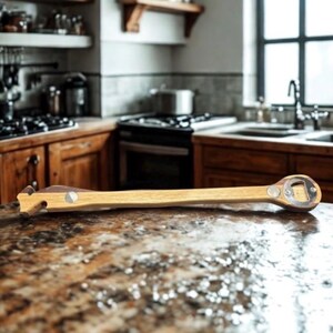 May include: A wooden bottle opener with a long handle and a metal bottle cap opener at the end. The handle is light brown with a dark brown accent and a silver rivet. The opener is lying on a speckled countertop in a kitchen.