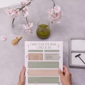 Op de afbeelding: Een werkblad voor de dagelijkse emotionele check-in met een bloemenontwerp wordt door een persoon vastgehouden. Het werkblad heeft secties voor stemming, lichamelijke sensaties en dagelijkse doelen. Een glas groene drank en kersenbloesems staan op de achtergrond.