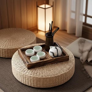 May include: A serene tea set arrangement on a woven mat. The set includes a wooden tray with small white teacups, a teapot, and a container with tea utensils. A woven floor cushion and a lit lamp are in the background.