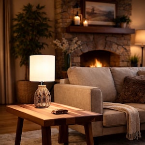 May include: A small table lamp with a woven base and a white lampshade sits on a wooden side table. The lamp is turned on, casting a warm glow. The table is next to a beige sofa and a fireplace.