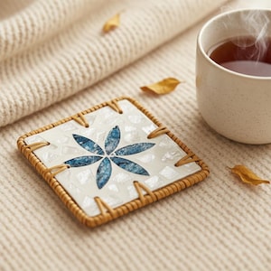 May include: Square coaster with a woven border and a mosaic design featuring a blue flower on a white background. A cup of tea sits next to the coaster, with steam rising from the top. The coaster is surrounded by a beige knitted fabric.