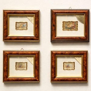 May include: Four framed miniature artworks with ornate, dark wood frames. Each piece features a small, rectangular, gold-toned artwork with a detailed, colorful scene. A cream-colored mat surrounds each artwork.