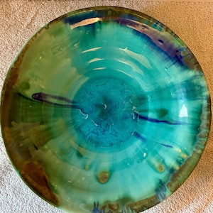 May include: A round ceramic bowl featuring a vibrant glaze with blended shades of turquoise, teal, and deep blue. The surface displays a glossy finish with organic, flowing colour transitions and darker brown accents along the outer rim edge.